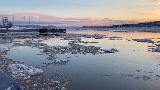 Ice floating on the St. Lawrence river seen during a partly cloudy late winter dawn, with row of flags on pier in the background, Quebec City, Quebec, Canada