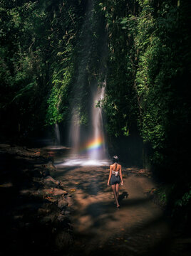 A young woman walks toward a tropical waterfall with a rainbow in a lush jungle at Suwat Waterfall in Ubud, Bali, Indonesia.
