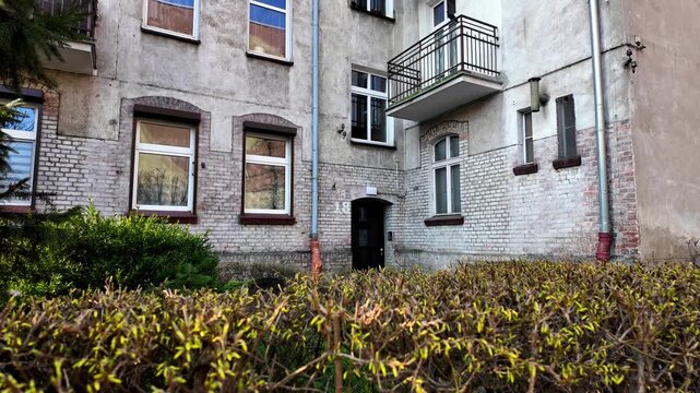 Exterior of an old white brick tenement building with entrance door, windows, and balcony, viewed behind a green hedge, featuring weathered facade and urban architectural details.