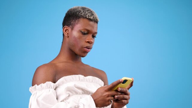 Young african american non binary person concentrating while typing a message on their smartphone against a solid blue studio background