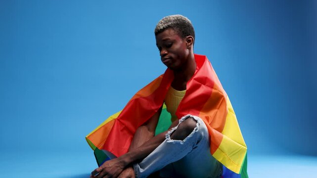 Proud african american man with dyed hair sitting on the floor wrapped in a rainbow flag. A symbol of the LGBTQIA plus community and pride month