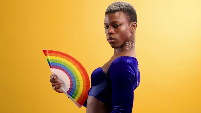 Confident Black non binary person holding a rainbow fan, turning and looking at the camera against a vibrant yellow studio background