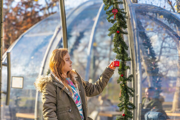 A happy young girl in a warm parka looking at a red Christmas ornament on a festive green garland outside a transparent dining dome in a sunny winter park in Budapest, Hungary today © Igor