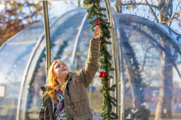 A happy young girl in a warm parka looking at a red Christmas ornament on a festive green garland outside a transparent dining dome in a sunny winter park in Budapest, Hungary today © Igor