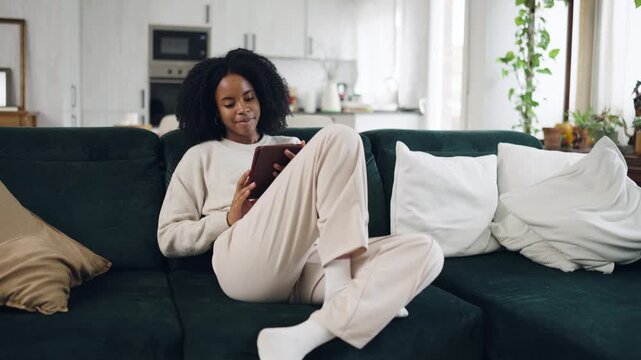Relaxed young woman with curly hair sitting on a sofa in her modern living room. She is happily browsing the internet and shopping online