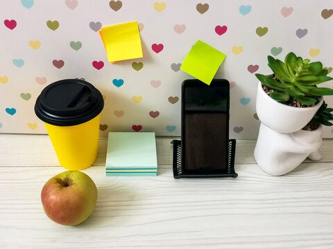 Yellow coffee cup, smartphone on stand, apple and sticky notes on white wooden desk with heart pattern wallpaper. succulent plant. Workspace for freelance, study, business, remote work