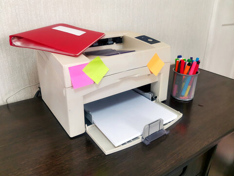 Beige printer with red folder and colorful sticky notes on dark wooden desk. Mesh holder with markers beside. Home office, business documentation, remote work workspace environment