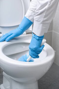 Person in blue gloves cleaning a toilet bowl with a brush. Close-up on hygiene and sanitation tasks in a modern bathroom. Bright, clean composition with white, blue, and grey colors.