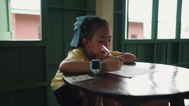 Asian girl focused on writing with a big bow on her head and a big watch on her wrist sitting next to a wooden table