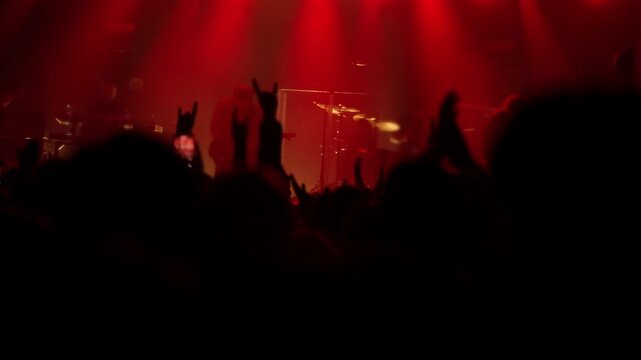 Excited crowd at a live rock concert or music festival, raising hands and making rock on gestures. The stage glows with intense red lights as the band performs