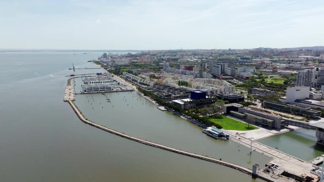 Aerial drone footage flying forward over the modern Parque das Nacoes waterfront in Lisbon, Portugal. Shows the marina, contemporary architecture, and the Tagus River on a sunny day