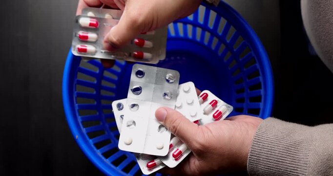 Detailed shot of hands tossing pharmaceutical blister packs into a blue garbage bin for medical negligence and drug waste themes.