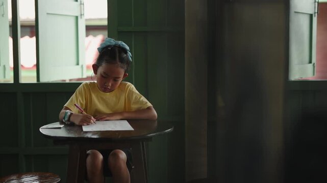 A little girl with a blue bow writes concentrated on a wooden table in a room with green windows and natural light