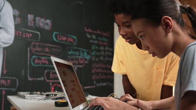 Happy african boy pointing at laptop screen while cute girl typing laptop coding or programming engineering system. Teacher teaching program while holding laptop at STEM technology class. Edification
