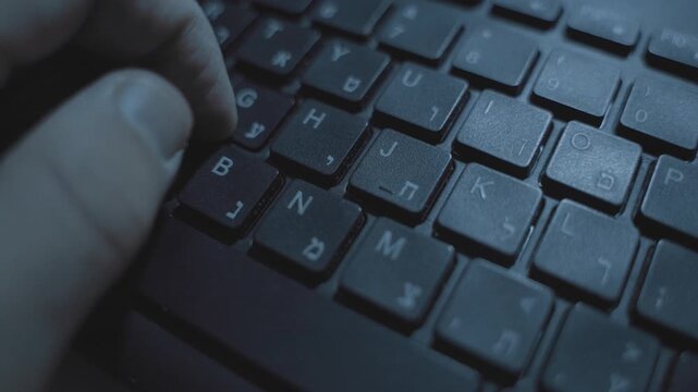 Hand typing on computer keyboard with Hebrew letters in dark office desk. Mysterious person working late at night clicking and pressing on keys. Hacker spy coding or creative writer at work business