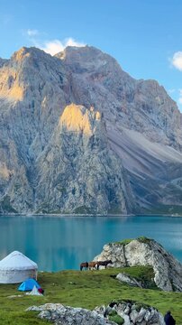 Video of tranquil blue Tian Tang Lake in Xinjiang Province, China, with mountains in the background as the sun rises over the mountain peaks. Horses are grazing nearby, and tents are pitched.