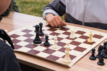 Close-up of two people playing chess outdoors on a wooden table. Strategy and competition concept. Hand of a player resting near the chessboard with plastic pieces © Kos