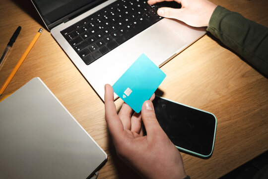 A person uses a debit card and a laptop for online transactions, with a smartphone and notebook on the wooden desk.