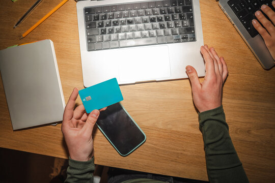 A person holds a debit card and a smartphone while using a laptop for online transactions.