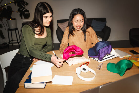 Two young women are studying together at a table, surrounded by books, notebooks, and colorful caps.