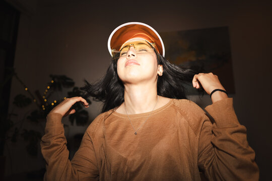 A young woman with dark hair wears a stylish visor and glasses, her eyes closed as she moves her head and hair, enjoying a moment of freedom.
