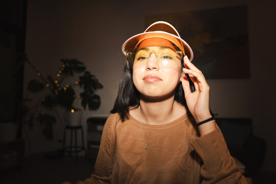 A young woman adjusts her stylish visor and glasses, posing indoors with a warm, inviting atmosphere.