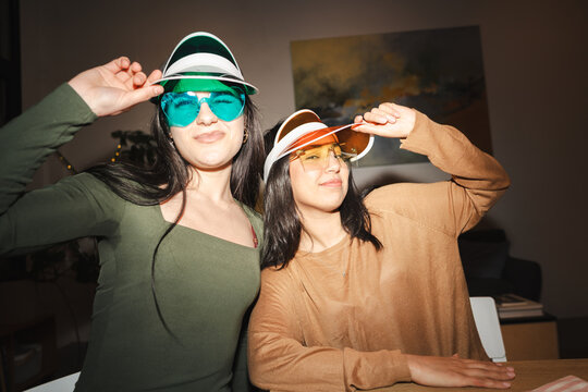 Two young women wearing colorful visors and heart-shaped sunglasses pose playfully indoors, enjoying a fun moment together.