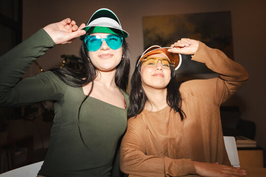 Two young women wearing colorful visors and oversized sunglasses pose playfully indoors, enjoying a fun and festive moment together.
