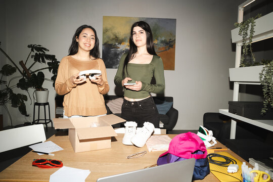 Two young women are unpacking a box of new items, including shoes and headphones, on a wooden table.
