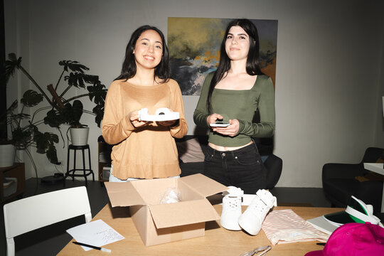 Two young women are unpacking a box of new white sneakers and other items in a room.