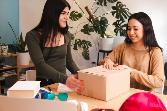 Two young women are packing a cardboard box with items on a wooden table, preparing for shipment.