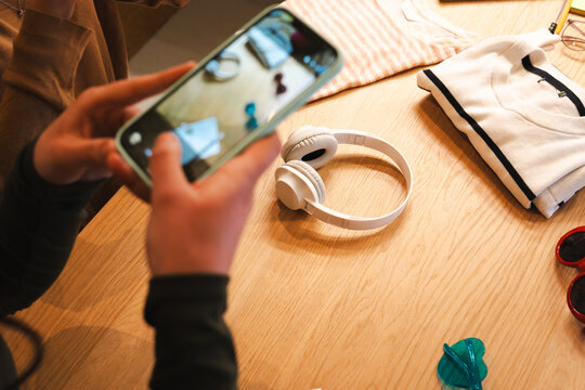 A person holds a smartphone, capturing a flat lay of clothing, headphones, and sunglasses on a wooden table.