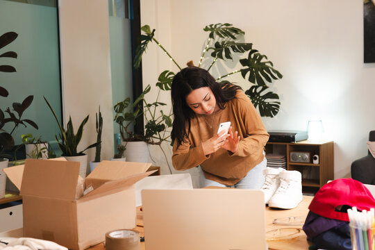 A young woman is using her smartphone to photograph items on a table, including an open box and a laptop.