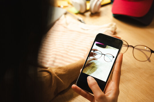A person holds a smartphone, capturing a close-up image of eyeglasses on a wooden surface. The phone screen displays the camera interface.