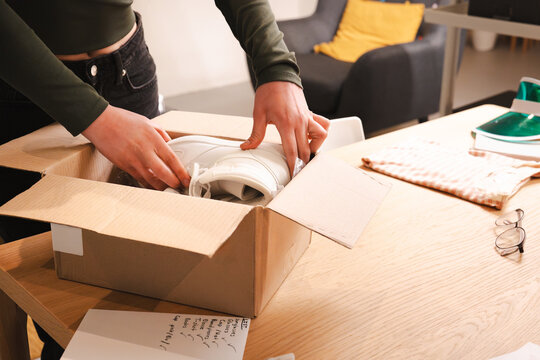 A person unpacks a new pair of white sneakers from a cardboard box on a wooden table, with a shopping list nearby.