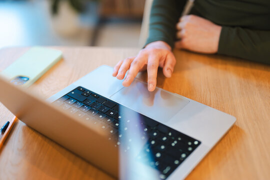A person's hand is using the touchpad of a laptop on a wooden desk. A mobile phone and a pen are also visible.