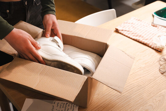 A person unpacks a new pair of white sneakers from a cardboard box on a wooden table, ready for a new adventure.