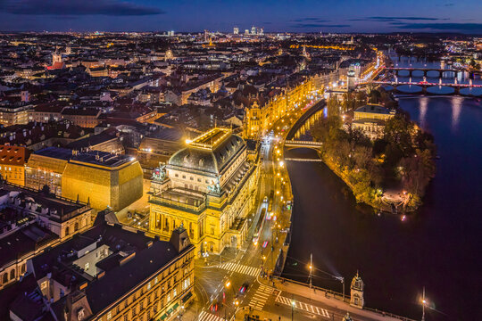 National Theatre in Prague stands illuminated along the embankment at dusk. The view shows the city skyline and river as lights reflect on the water.