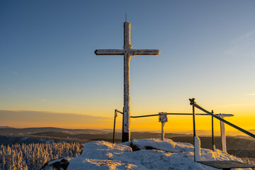 The cross stands on the top rock formation of Jizera Mountain. Snow covers the ground, and the sun sets behind distant hills. Trees are visible below the summit. © pyty