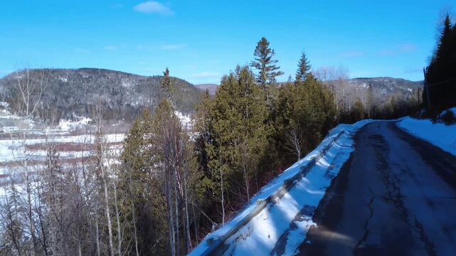 Cinematic drone shot of a winding winter road through a snow-covered forest and mountains near the frozen Restigouche River in Matapedia, Quebec. Crisp blue sky and pristine Canadian wilderness.
