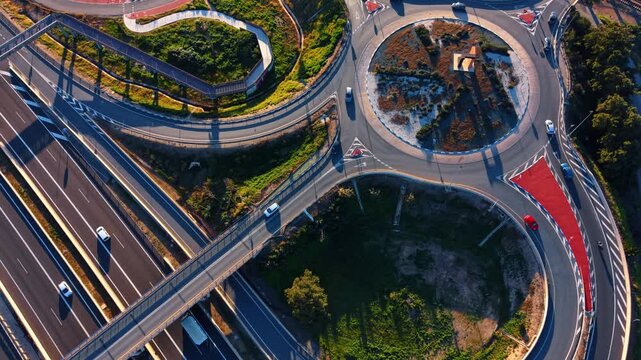 Many cars drive by the roundabout, trestles and highways at sunset. Modern traffic system from drone.