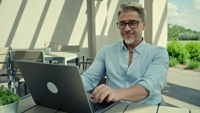 Businessman sitting at table in outdoor cafe, working on laptop and smiling. Happy successful mature man with gray hair and glasses remote managing business from home office on peaceful sunny day.