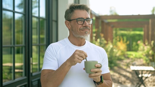 Mature man wearing eyeglasses and white t-shirt drinking coffee in mug, standing in backyard garden, looking away. Confident senior male smiling outdoor.