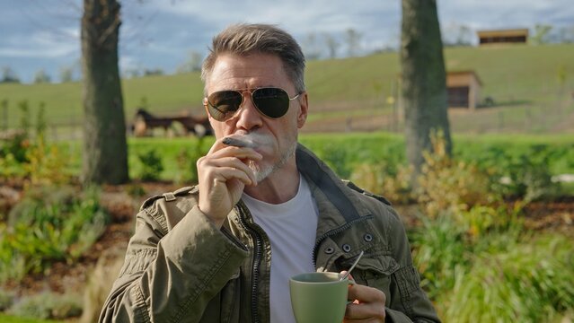 Mature farmer enjoying cigar and coffee on ranch, in front of rural landscape. Mid adult man with grey hair and sunglasses. Successful agricultural businessman on his land at morning.