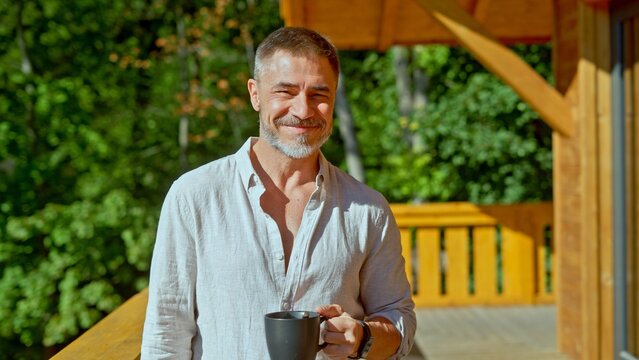 Portrait of middle aged man with gray hair in casual cloth outdoor on home balcony. Mid adult male starting the day with morning coffee. Spring or summer. Happy satisfied smile.