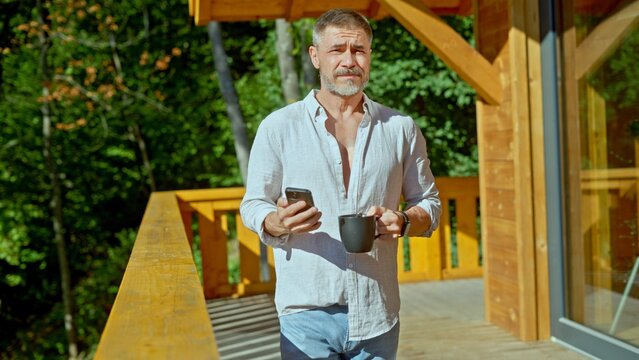 Businessman starting the day with morning coffee and checking phone. Portrait of middle aged man with gray hair in casual cloth outdoor on home balcony. Spring or summer.