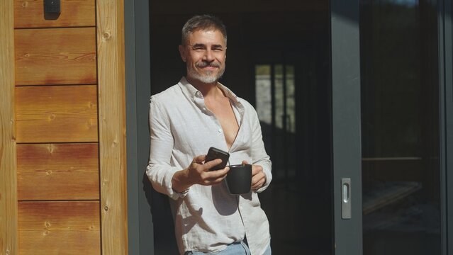 Businessman starting the day with morning coffee and checking phone. Portrait of middle aged man with gray hair in casual cloth outdoor on home balcony. Spring or summer. Happy satisfied smile.