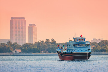 Fototapeta premium Traditional dhow boat cruising along Dubai Creek with city skyline and palm trees during a vibrant sunset