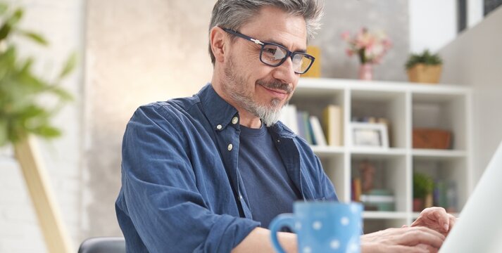 Happy man with laptop computer at home. Businessman working at desk in home office. Portrait of mature age, middle age, mid adult man, bearded, glasses, smiling, authentic look.