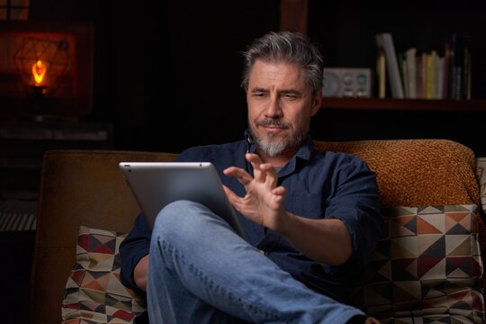Man sitting on couch at home with tablet computer. Businessman working in home office. Portrait of mature age, middle age, mid adult man, bearded, authentic look.